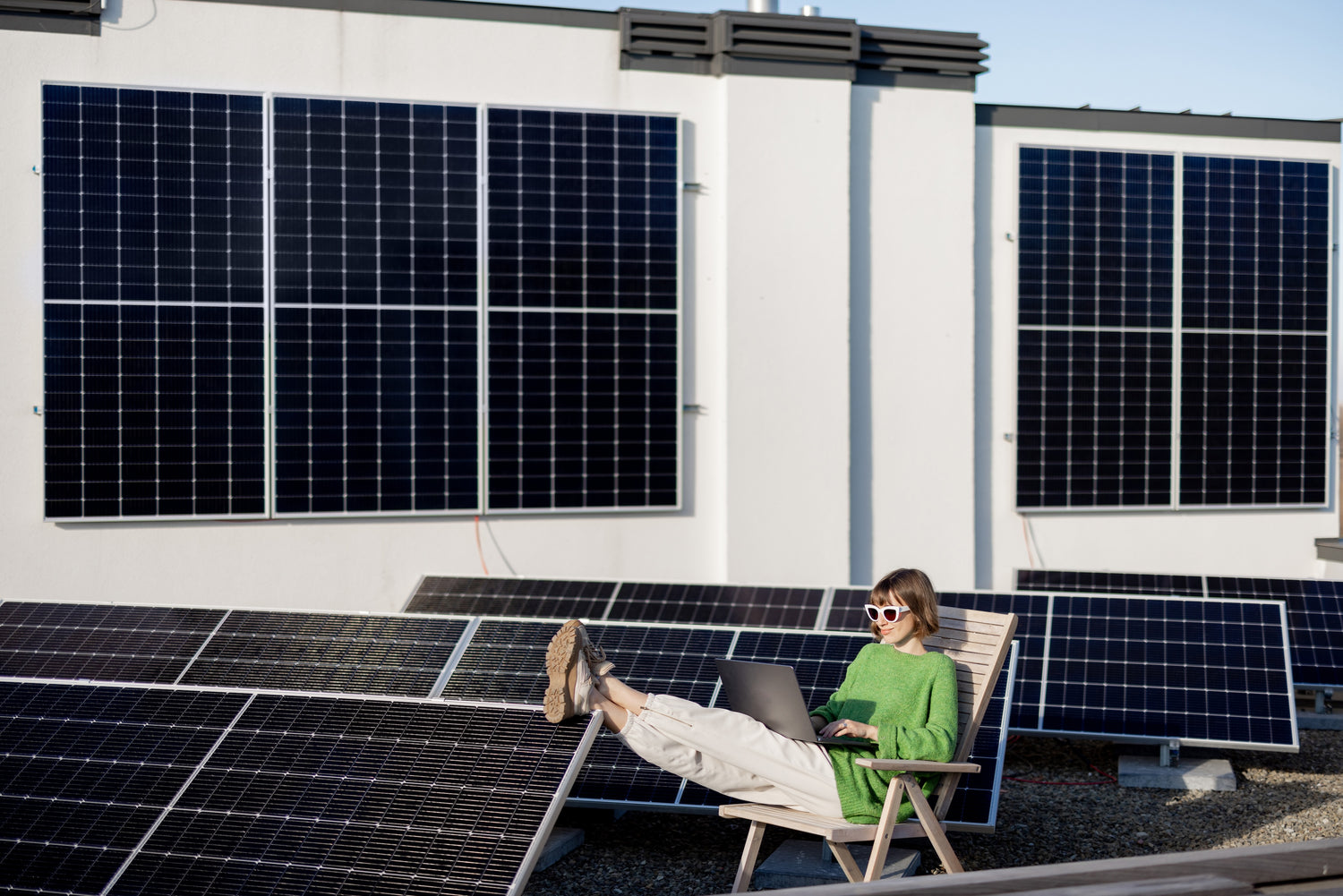 Il tetto di un'abitazioe civile tappezzato di pannelli fotovoltaici di diverse dimensiuoni. Una ragazza sta leggendo seduta su una sedia di legno con pantaloni verdi e bianchi e occhiali.