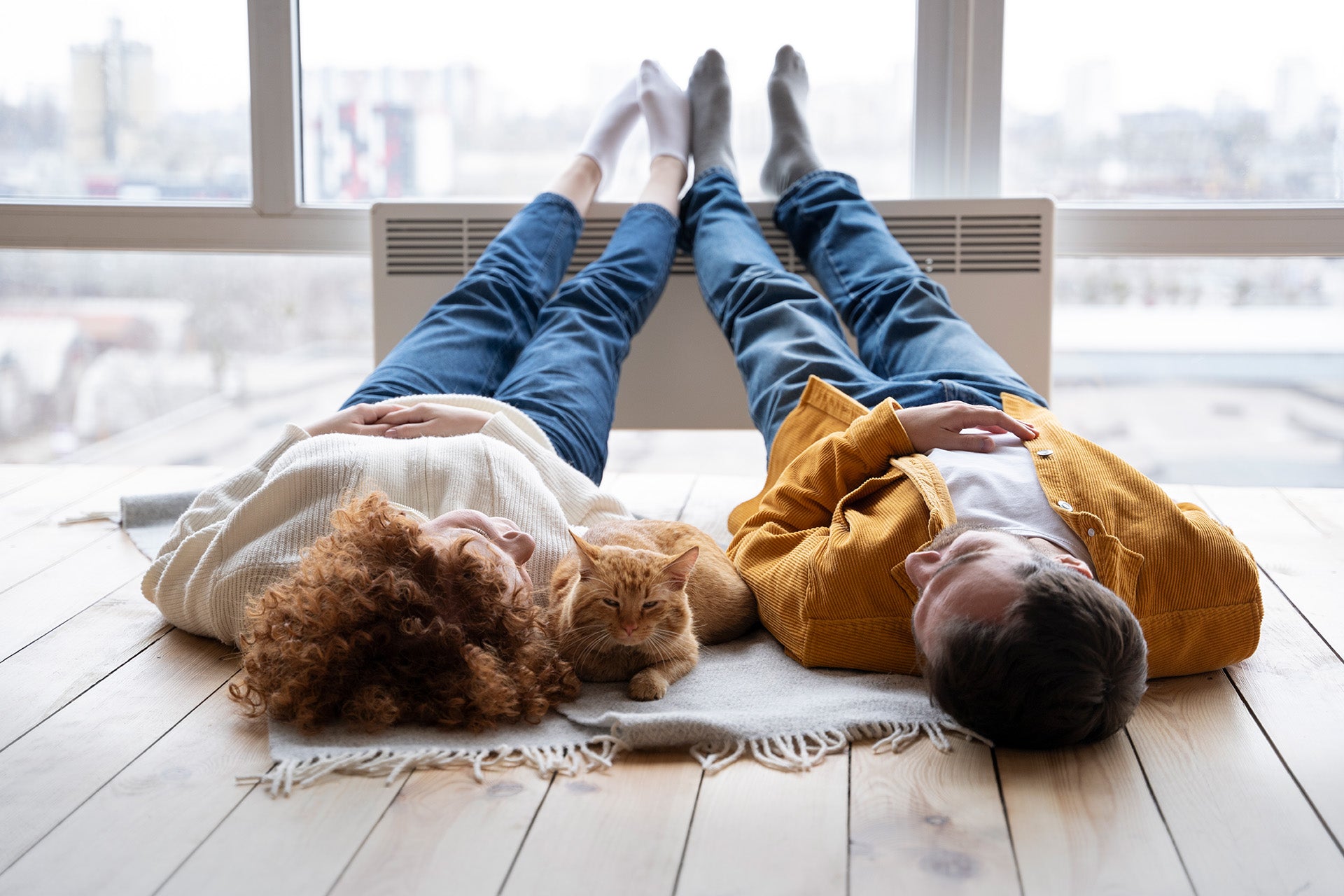 Una coppia sdraiata su pavimento di legno con piedi appoggiati su uno ventilconvettore. Sullo sfondo una vetrata con vista su una città. 